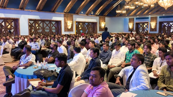 A large group of men attending a business meeting or conference in a hotel ballroom, with a speaker visible in the background.