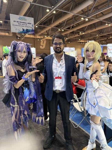 A man in a suit poses with two cosplayers at a trade fair, with an event banner in the background.