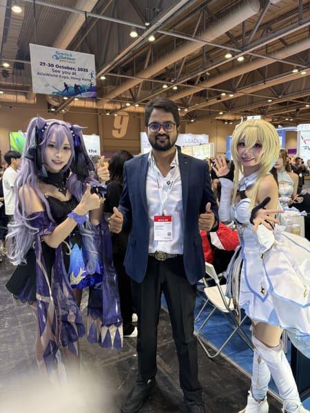 Man in suit with two cosplayers at a trade fair in Hong Kong, smiling and giving thumbs up.