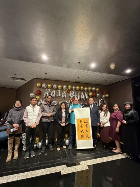 Group of nine people posing with a scroll in front of birthday balloons, celebrating an event indoors.
