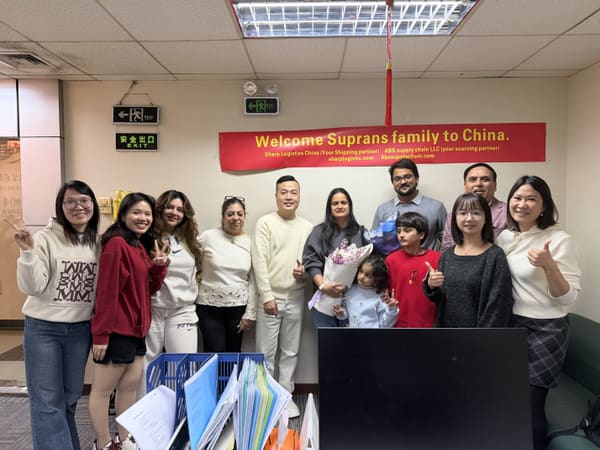 Diverse group of 13 people, including children, smiling in an office with a "Welcome Suprans family to China" banner.