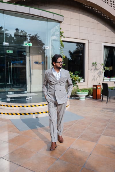 A man in a light grey double-breasted suit and brown shoes walks outside a Radisson hotel, looking to his right.
